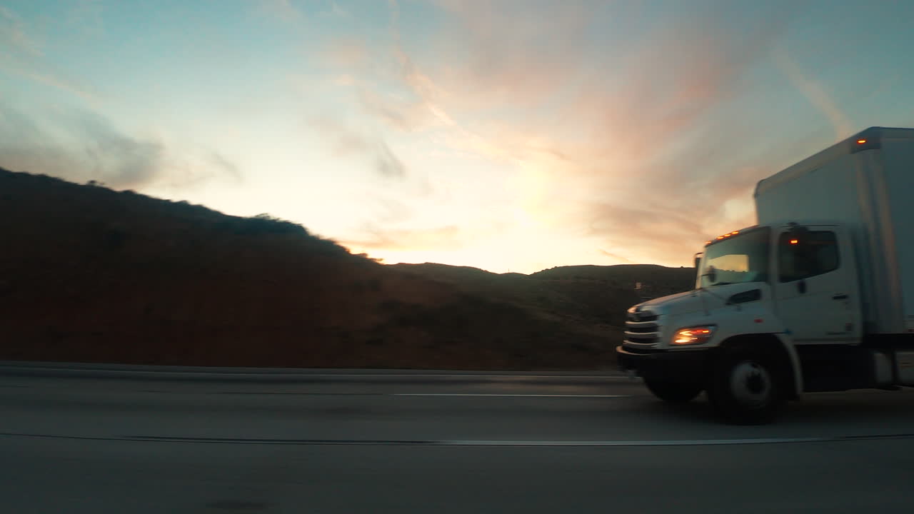 Caminhões viajando nas rodovias juntamente com outros veículos passando, com belas montanhas durante o pôr do sol - Plano Aberto