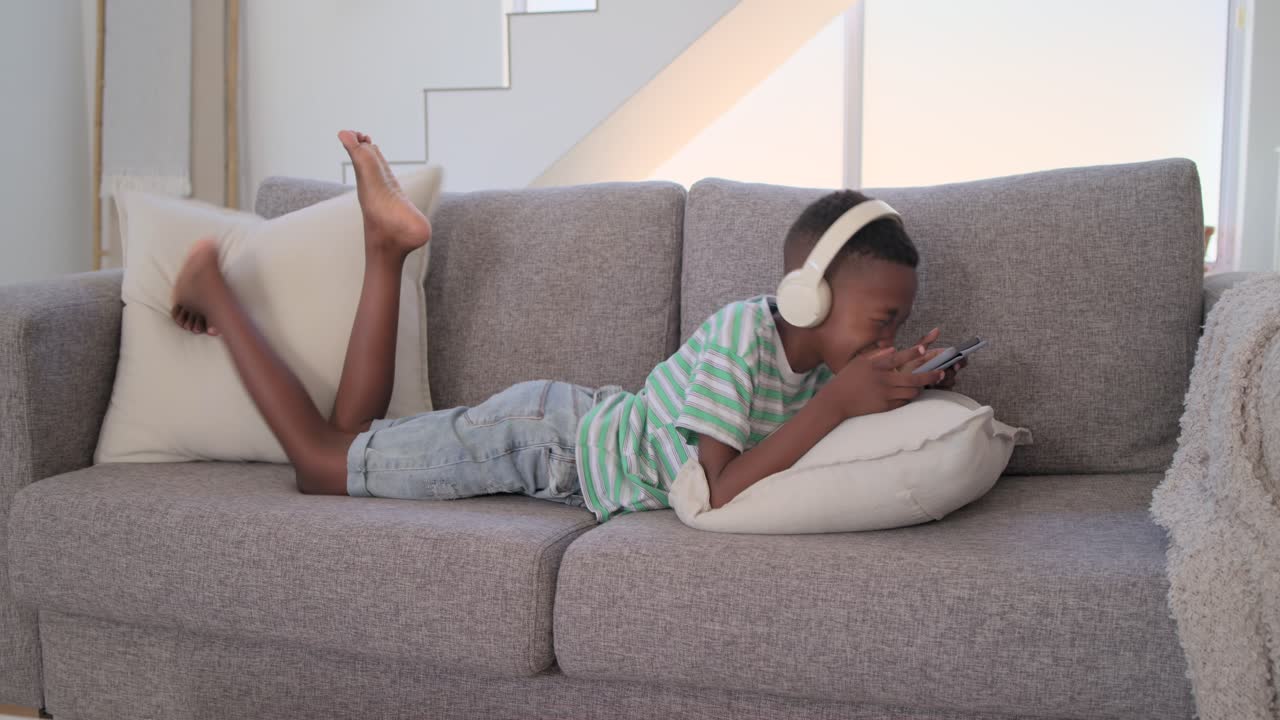 Afroamerikansk ung person ligger på en grå sofa i stuen, holder en telefon, har hovedtelefoner på og griner.