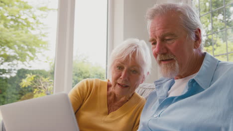 retired senior couple sitting on sofa at home using laptop online shopping or booking holiday