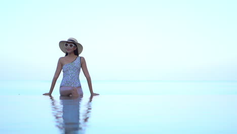 Asian-Lady-sitting-at-the-edge-of-the-infinity-pool-leaning-on-arms-with-crossed-legs-under-the-water-slow-motion-static,-copy-space-template