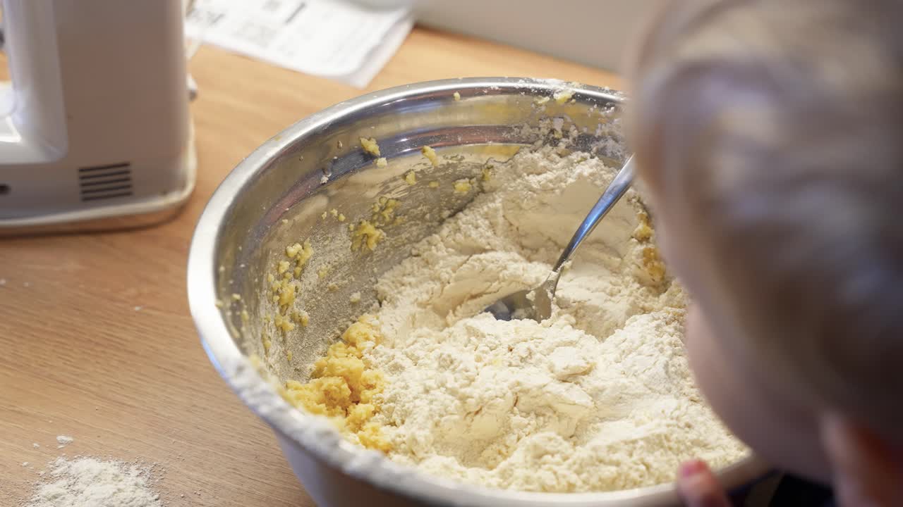 Et nærbilde av et lite barn som elter deig i en bolle av rustfritt stål fremhever en praktisk bakeaktivitet på et hjemmekjøkken. Mykt naturlig lys forsterker den hjemlige scenen.