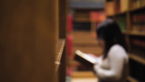 Sliding-left-shot-behind-a-bookshelf,-sharp-foreground-with-books,-and-blurred-figure-of-a-woman-holding-a-book-and-turning-a-page