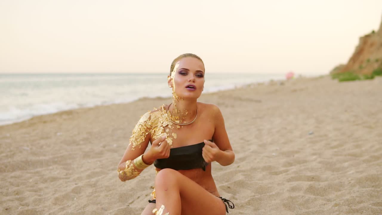Young model with professional golden art makeup lying on the beach looking in the camera and flirting with the viewer, pouring