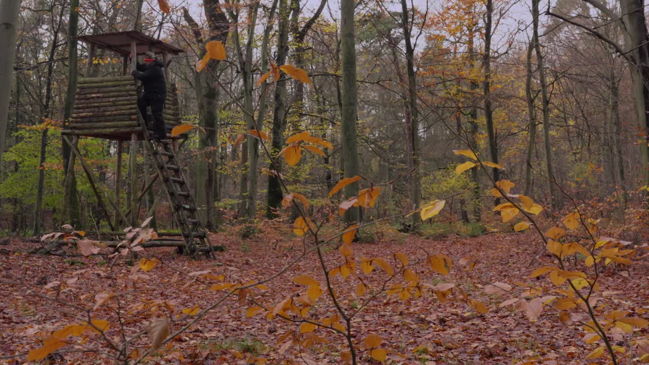 Campo lungo di un cacciatore in una spessa tuta invernale che scende da un'altana, portando un fucile da caccia con silenziatore in spalla.