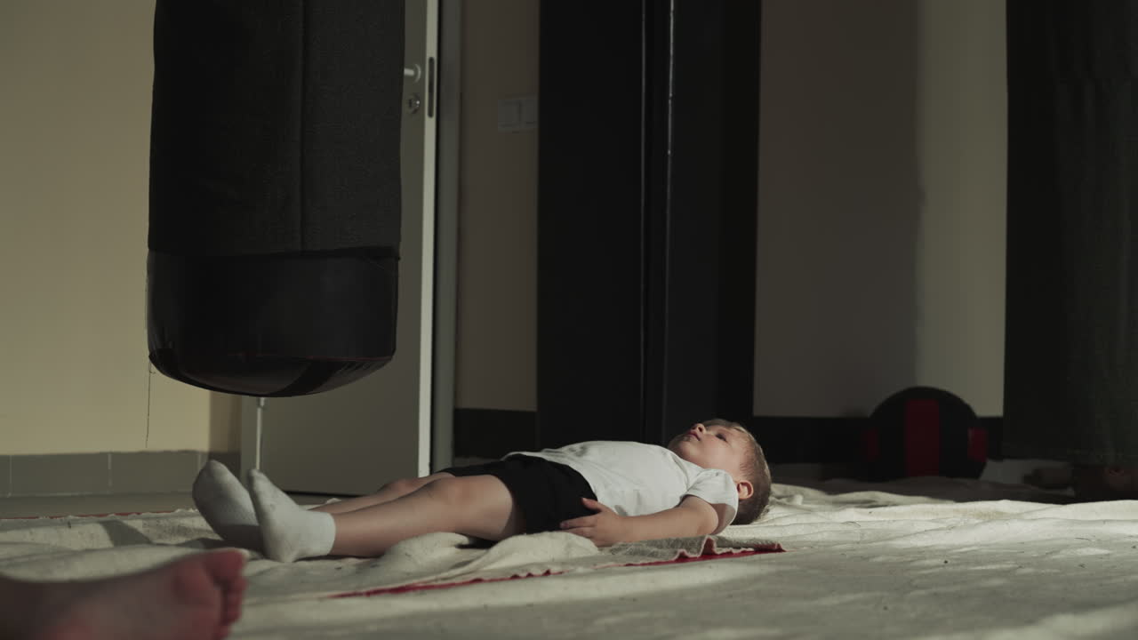 아이는 스포츠 센터에서 싱 봉투 근처에 휴식을 취합니다. 어린 소년은 어린이 체육관에서 무술 연습을 한 후 느린 동작으로 누워 있습니다. 유아는 바닥 개에 긴장을 풀고 있습니다.