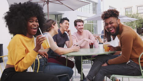 six millennial friends sitting in the street outside a cafe talking, turn to camera smiling