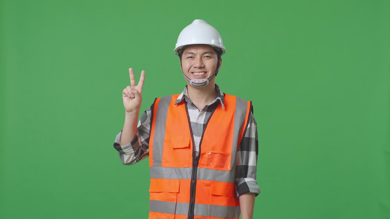 Asian Male Engineer With Safety Helmet Smiling And Showing Peace Gesture While Standing In The Green Screen Background Studio