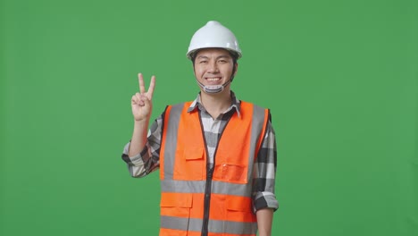 Asian Male Engineer With Safety Helmet Smiling And Showing Peace Gesture While Standing In The Green Screen Background Studio
