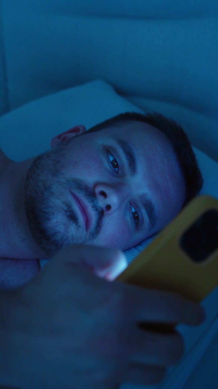 Uomo sdraiato sveglio a letto di notte che usa il suo smartphone, suggerendo insonnia, mancanza di sonno, dipendenza dallo schermo e uso notturno del telefono cellulare