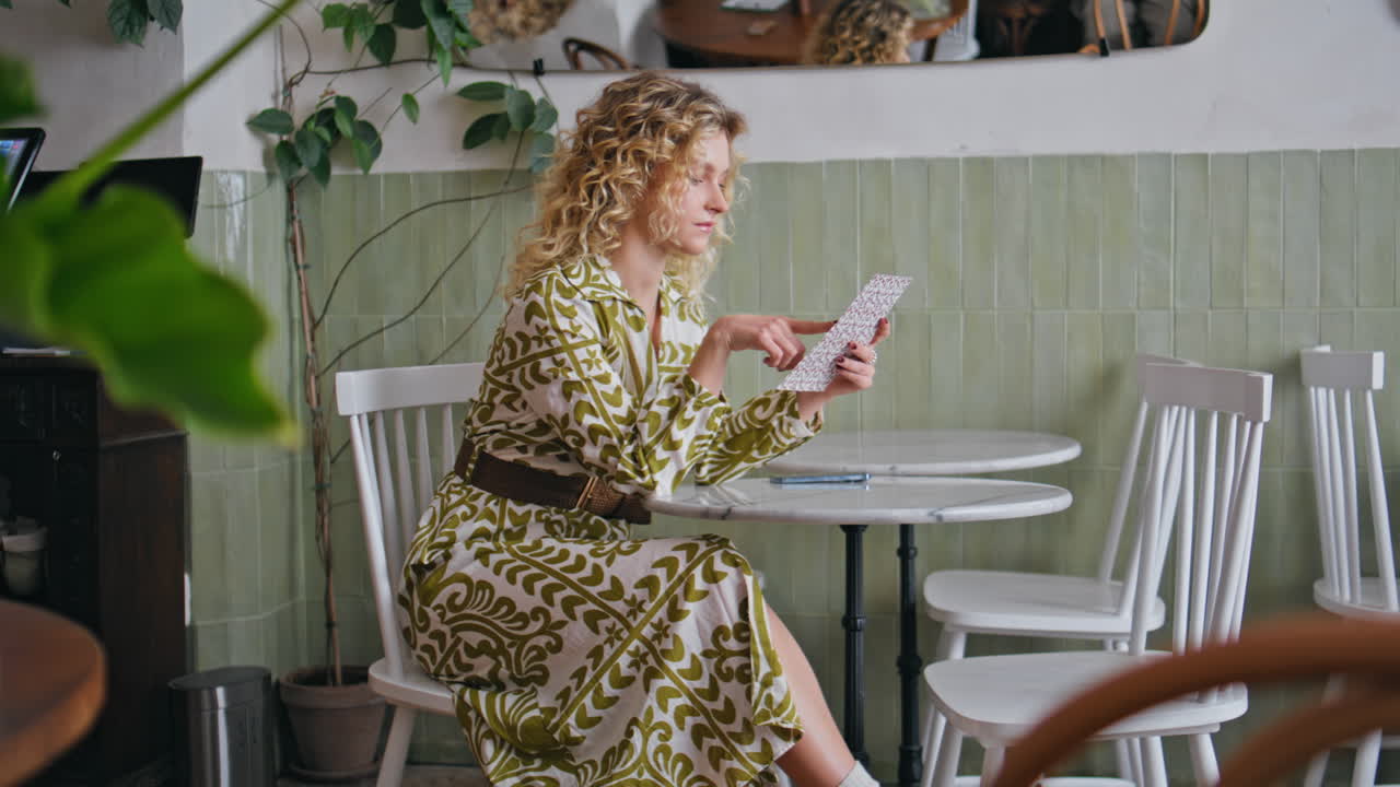 Relaxed lady reading menu in calm cafe. Pretty curly-haired woman choosing dish