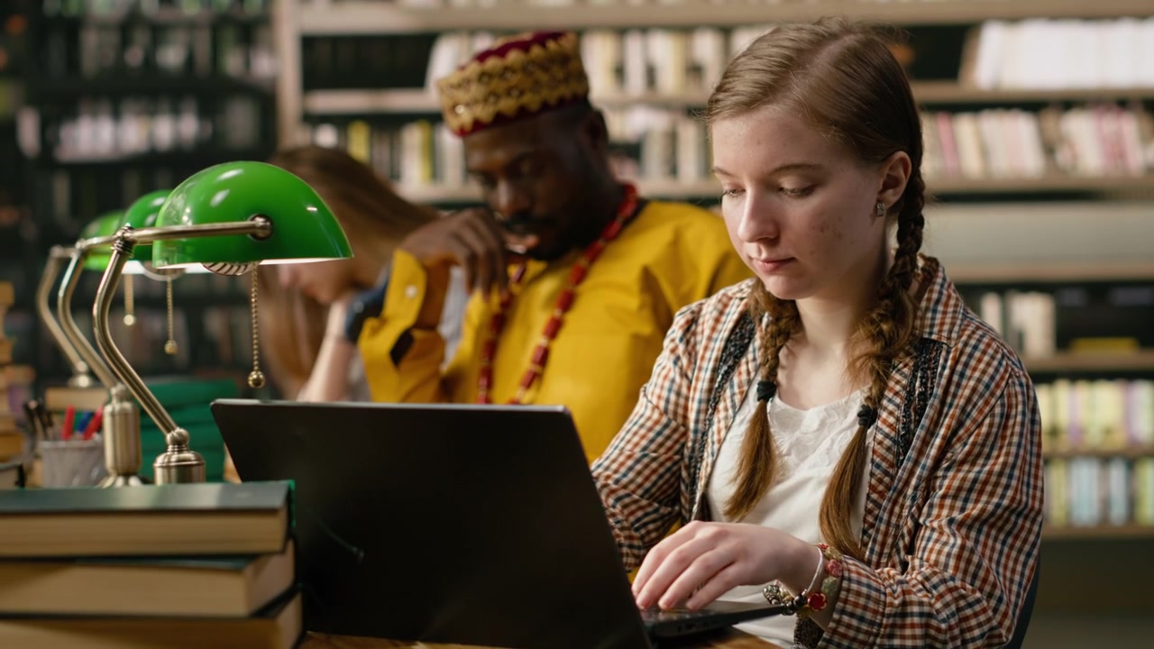 Student typing on laptop and taking notes beside open book at busy study table