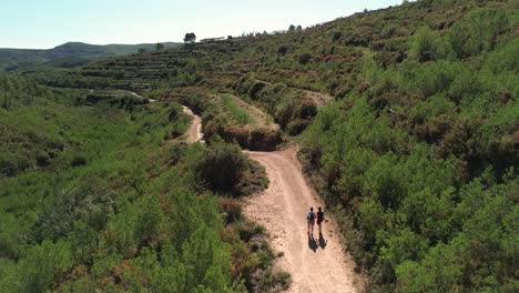 una fotografía de un avión no tripulado de dos chicas morenas caminando por un camino de tierra rodeado de coníferas, almendros y vegetación.