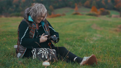 mujer gorda mayor sonriendo y mirando su cámara mientras está sentada en la hierba en la naturaleza rodeada de coloridos árboles de hojas naranjas y vegetación