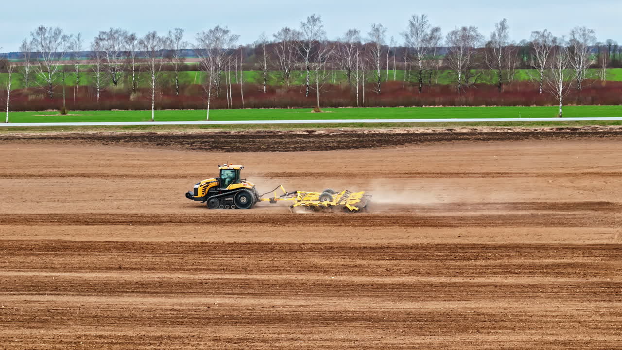 A smooth aerial parallax tracking shot follows a powerful Challenger tracked tractor as it pulls a cultivator to prepare the soil for spring planting on a large farm in Latvia.