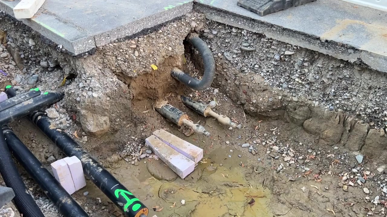 Open sleuf met zichtbare zwart omhulde stadsverwarmingsbuizen, golfde kabelgoten en isolatieblokken in natte, grindachtige grond. Werkzaamheden aan nuts- en energie-infrastructuur in een Duitse wijk