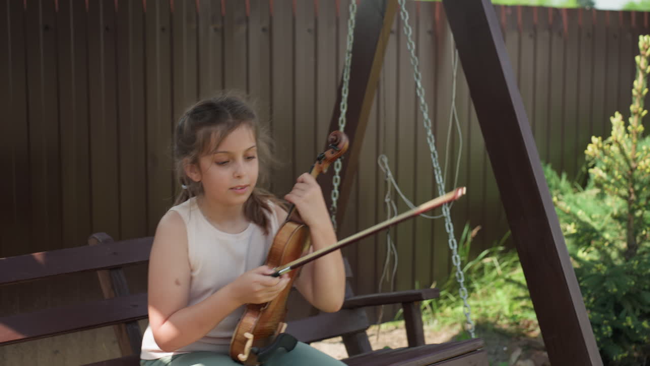 Young Caucasian Girl Practicing Violin On Swing, Sunlit Backyard Bench Scenes Show Tuning Strings, Adjusting Bow, Rehearsing Melodies, Focused Expression, Relaxed Summer Atmosphere, Wooden Swing