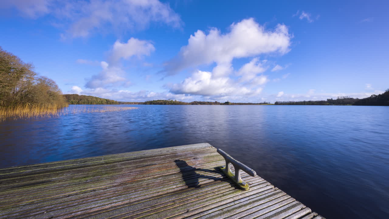 Time-lapse di un molo in legno sul lago in primo piano e un'isola con rovine di castello e foresta in lontananza, in una giornata nuvolosa e soleggiata a Lough Key nella contea di Roscommon in Irlanda.