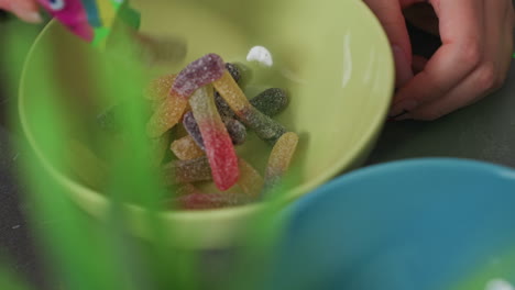 close up colorful gummy candies in yellow bowl with hand pouring candies from green pack, vibrant mix of red, yellow, orange, purple candies covered in sugar, casual indoor snack scene