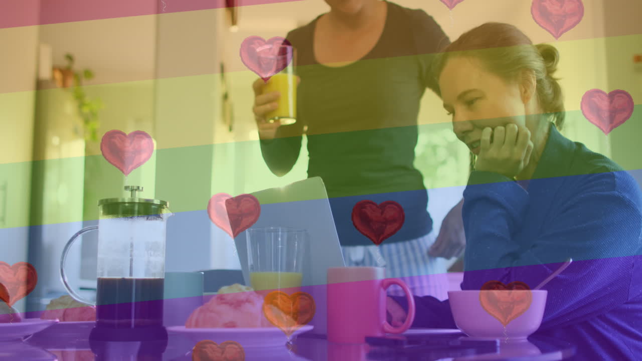 Animation of heart emojis and rainbow flag over caucasian female couple at home