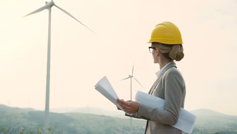 caucasian woman engineer wearing a helmet watching some blueprints at wind station of renewable energy