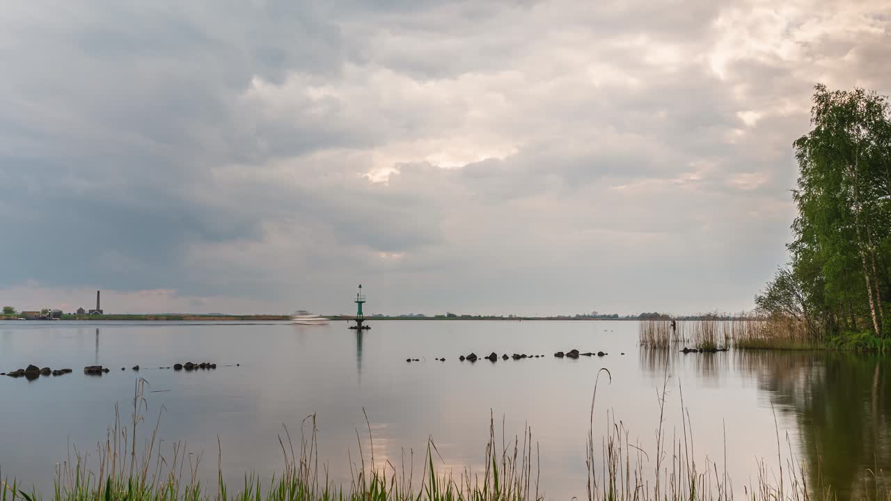 Lakeside timelapse with cloudy sky, moving boat, lighthouse, reeds and distant an old steam pumping station silhouette at dusk. Timelapse.