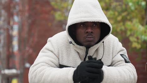 thoughtful young man cracks knuckles in white hoodie outdoors, resting on iron bar, blurred background features trees and buildings, creating a contemplative mood in a winter urban setting