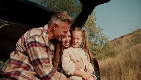 Close-up-shot-of-a-happy-middle-aged-brunette-man-with-a-little-gray-hair-talking-and-hugging-his-wife,-the-brunette-girl-in-a-green-checkered-shirt,-and-a-little-blonde-daughter-Sitting-in-the-open-trunk-of-a-car-on-his-picnic-Far-from-the-city