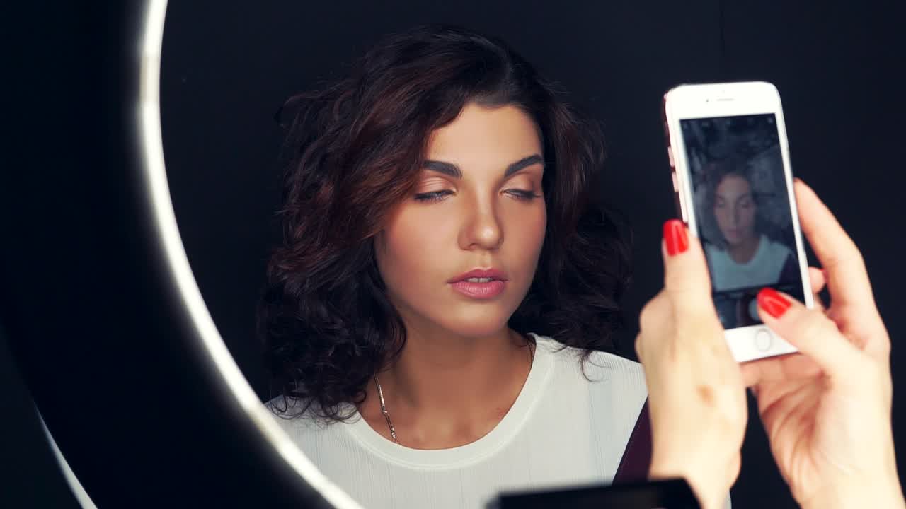fotografía de cerca de una mujer joven y hermosa siendo fotografiada con un teléfono móvil frente a la lámpara del anillo. modelo atractivo con