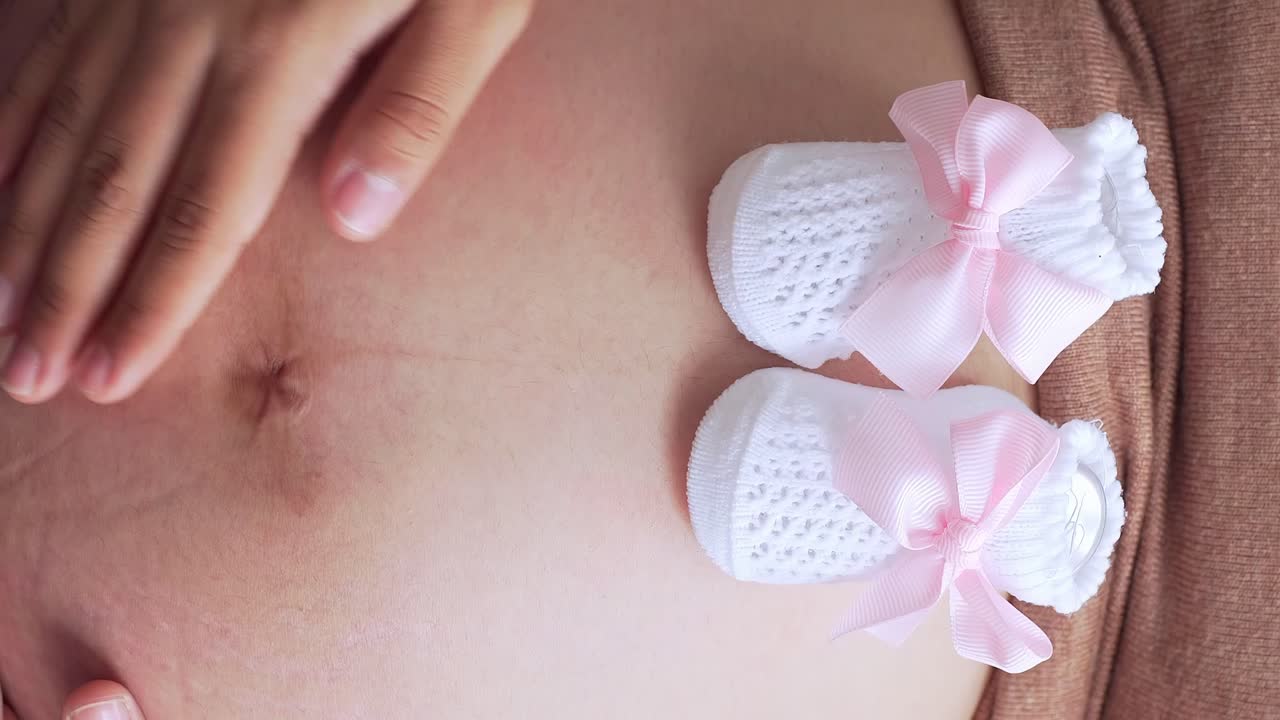 Malé růžové botičky na břiše těhotné ženy, detailní záběr, vertikální.