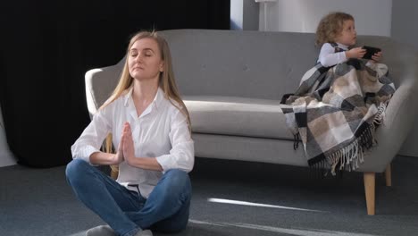 The Young Mother is Doing Yoga and the Child is Sitting on the Sofa Relaxation at Home the Young Mother is Doing Yoga and the Child is Sitting on the Sofa Relaxation at Home