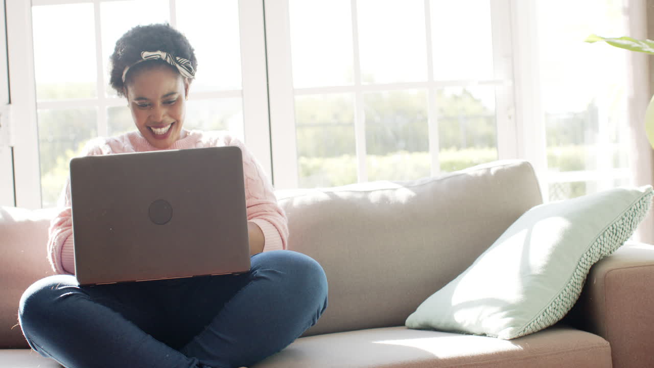 African American woman laughs while using a laptop on a sofa at home with copy space