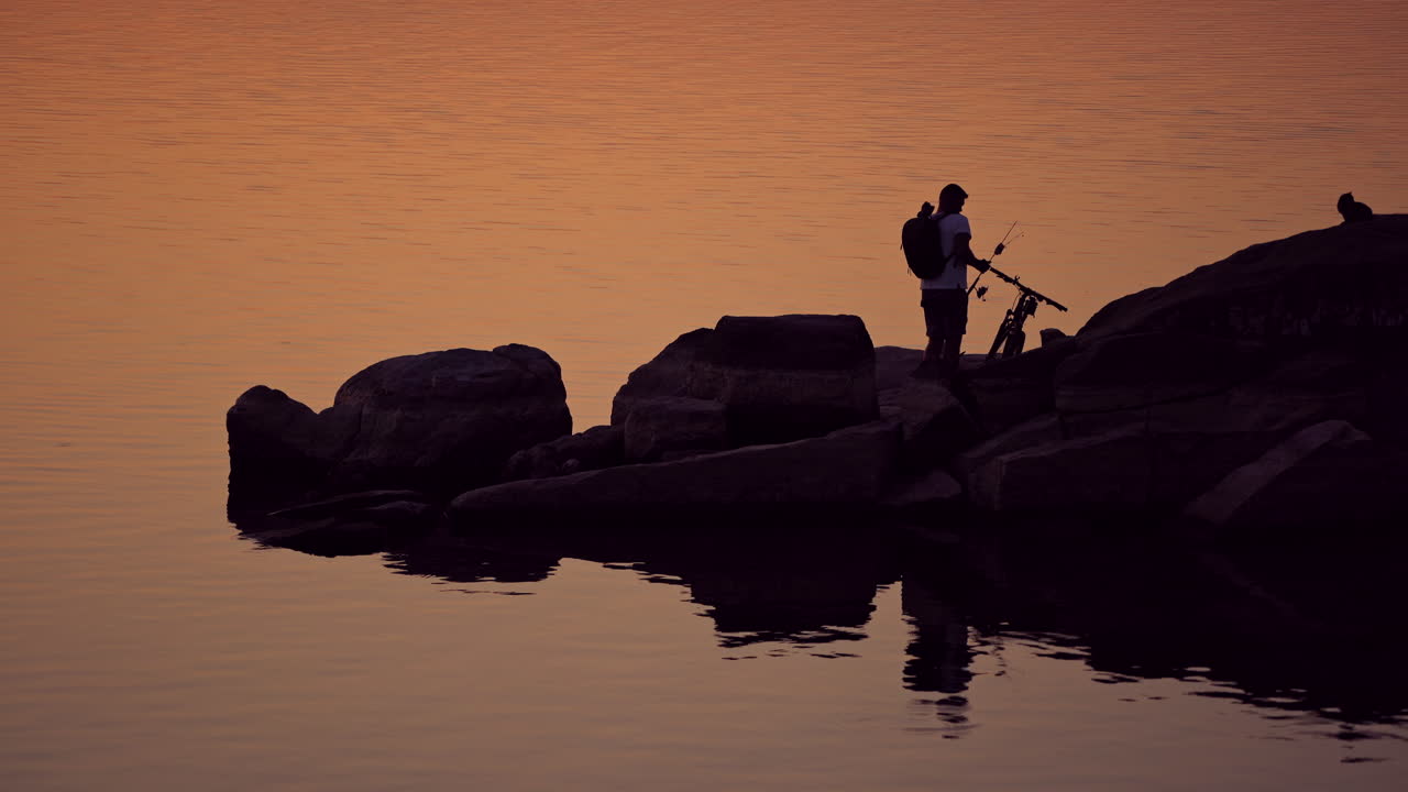 Man with fishing rods near his bike on the evening water background. Silhouette of a fisherman with rucksack is gathering home standing on stony rocks over water.