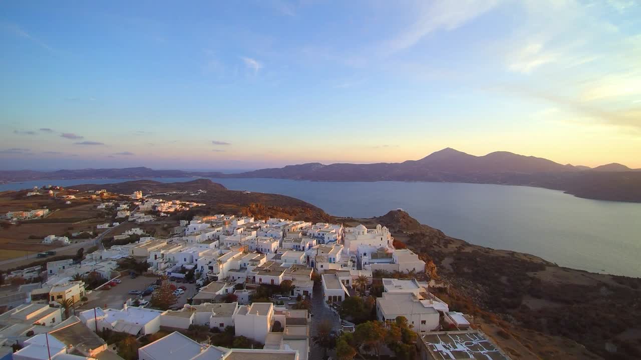 Aerial drone shot of peaceful coastal village with white buildings on hillside, calm sea and mountains in background, glowing under soft sunset light in warm serene Mediterranean setting