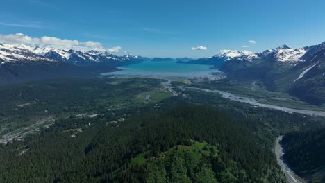 Dense-Pine-Forest-And-Calm-Blue-Lake-Between-Snowy-Mountains-In-Alaska-On-A-Sunny-Day