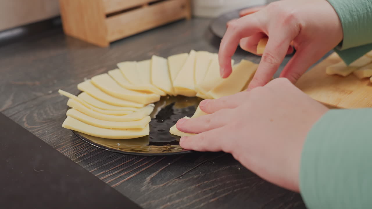 close up of woman taking sliced yellow cheese from wooden cutting board and arranging it neatly on black ceramic plate over dark wood table in kitchen, creating circular cheese presentation for meal