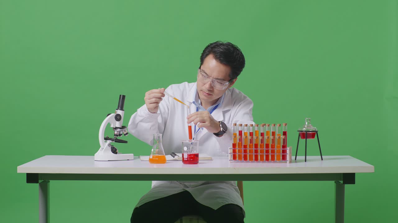 아시아인 과학자가 시험관 실험을 하고 노트북에 글을 쓰면서 초록색 스크린의 백그라운드 실험실에서 현미경으로 테이블에서 일하고 있다.