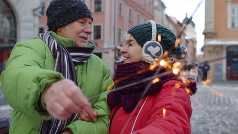 Pareja-Mayor-Sosteniendo-Luces-De-Bengala-Disfrutando-De-La-Víspera-De-Navidad,-Besándose-En-La-Ciudad-De-Invierno