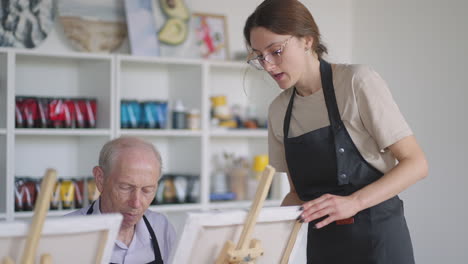 a female teacher shows a retired man how to draw a picture with paints and a brush at courses for the elderly. a senior man draws a picture to a group of pensioners