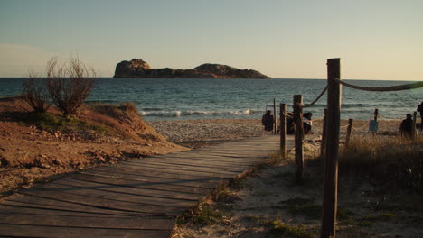 Pasarela-De-Madera-En-La-Playa-Con-Gente-Frente-A-La-Isla,-Puesta-De-Sol,-Porto-Tramatzu