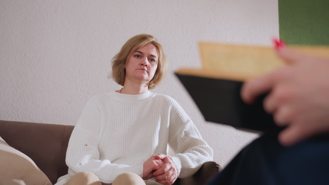 Participant sits attentively on sofa in white sweater, leaning slightly forward with focused expression while trying to see pages therapist is flipping through during calm therapy session
