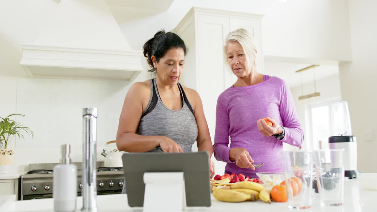 Dua wanita senior yang bahagia dan beragam sedang menyiapkan buah-buahan menggunakan tablet di dapur, gerak lambat