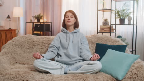 mujer joven meditando sentada en posición de loto en el sofá mirando tranquila con los ojos cerrados en casa