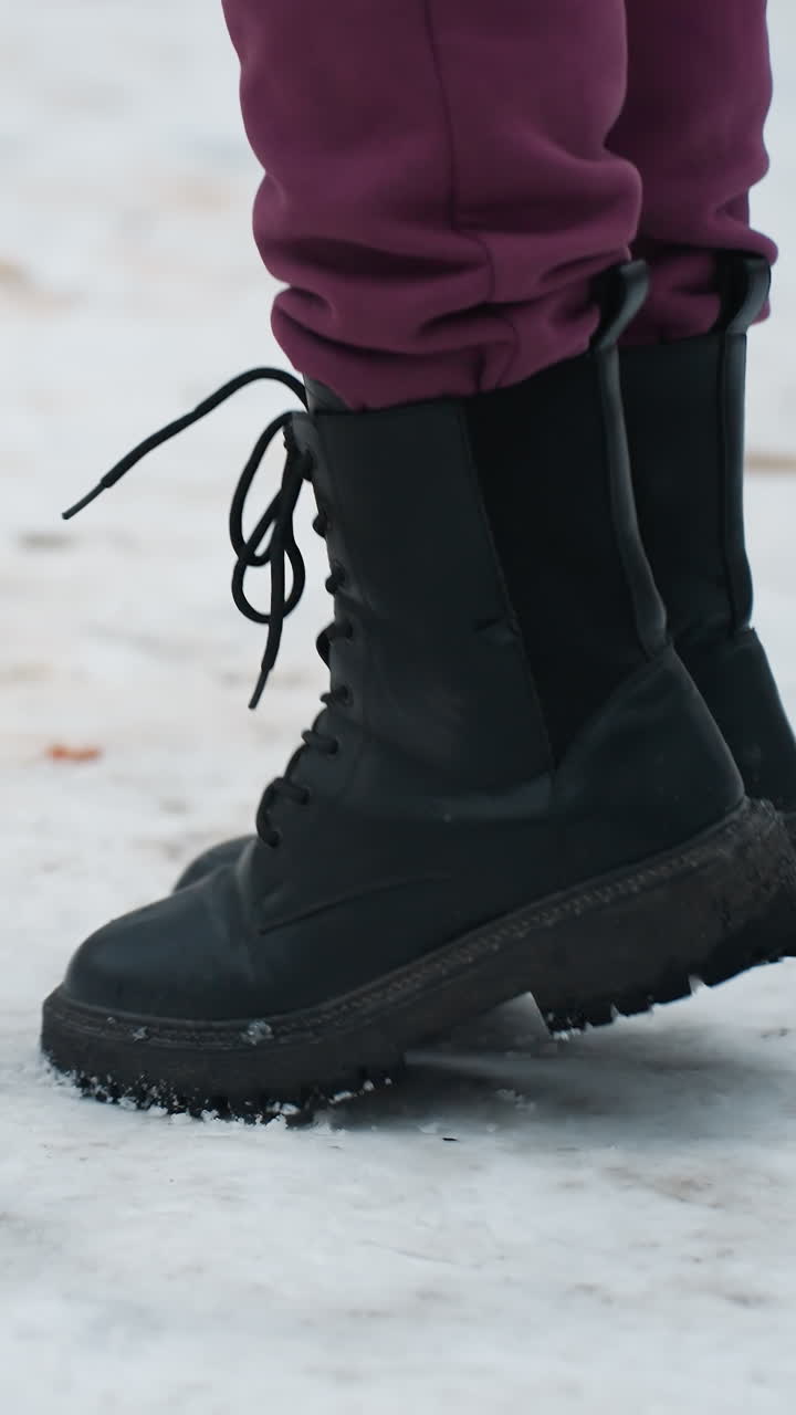 vista lateral de un individuo con botas negras saltando de derecha a izquierda en un terreno cubierto de nieve con follaje seco esparcido, la escena de invierno destaca el movimiento, el calzado y la textura del terreno helado