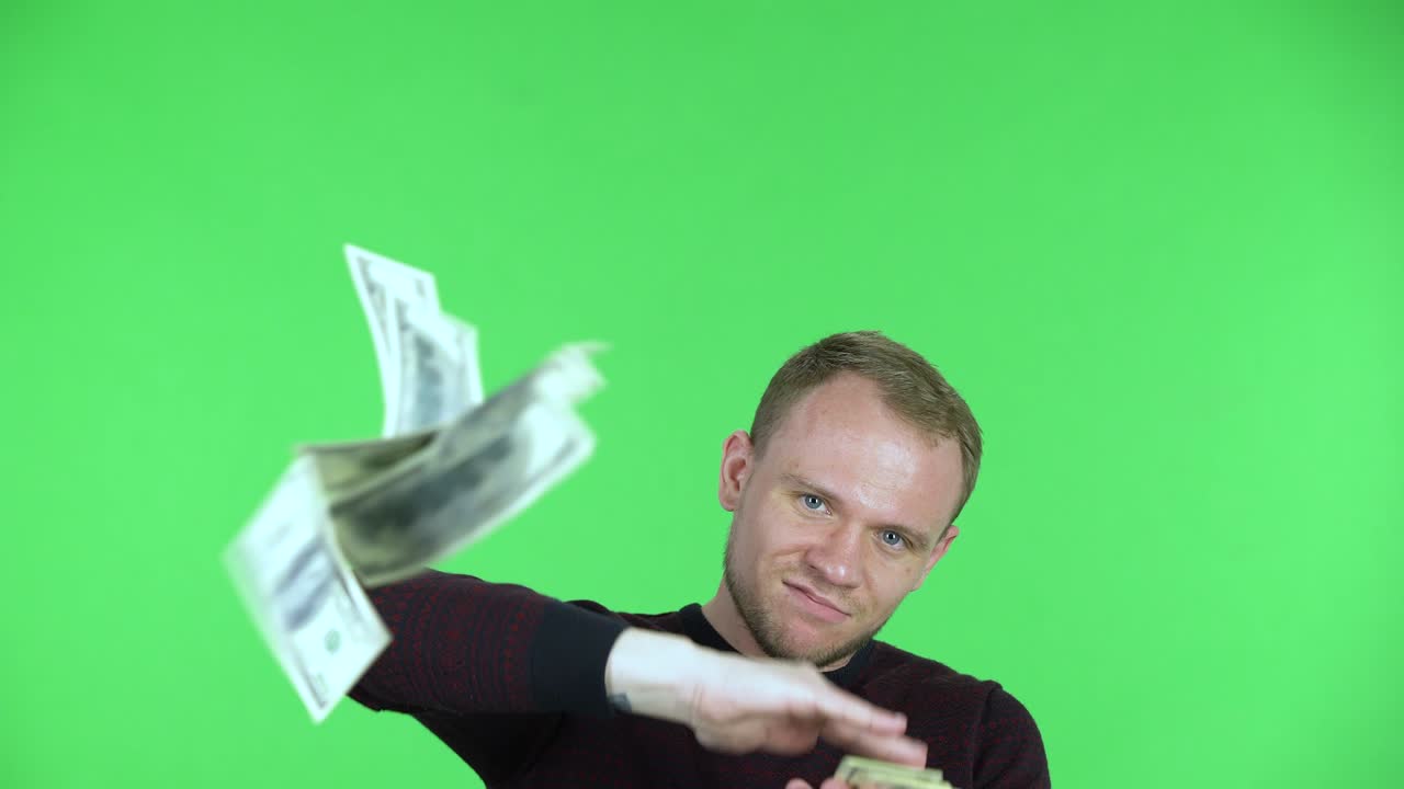 Portrait of a middle aged men scattering dollar bills and showing off his wealth. Unshaven male in a black red sweater posing on a green screen in the studio. Close up