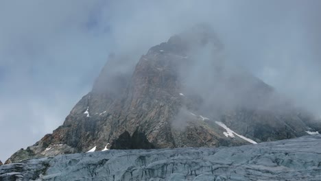 Drone-Volando-Sobre-El-Glaciar-Fellaria-Con-Majestuosa-Montaña-Rocosa-Envuelta-En-Nubes-En-El-Fondo-En-Un-Día-Soleado-De-Verano,-Valmalenco-En-Italia