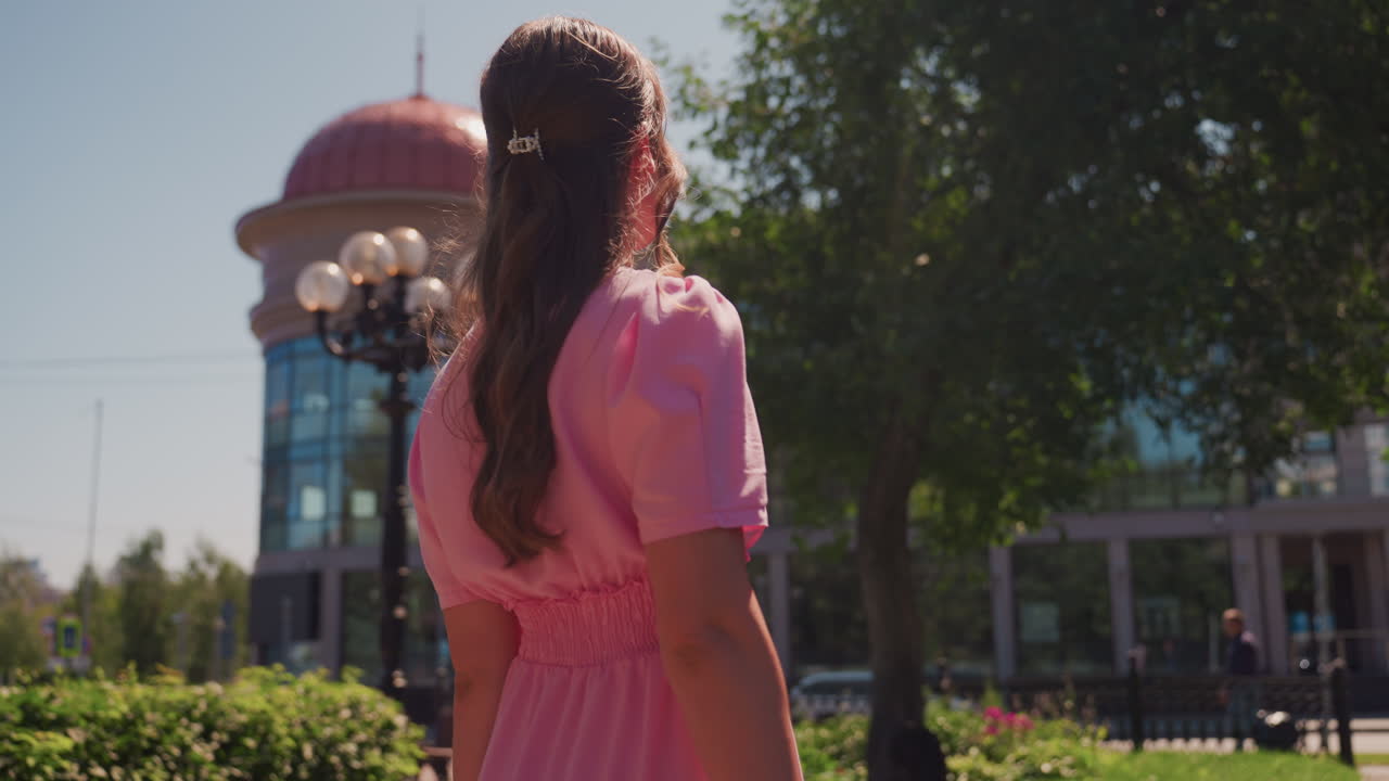 joven con vestido rosa explorando una plaza moderna, cúpula de cristal reflectante y fachada de edificio, setos y macizos de flores bien cuidados, sombra de árboles altos, detalle de farola, girando la cabeza con una mirada curiosa