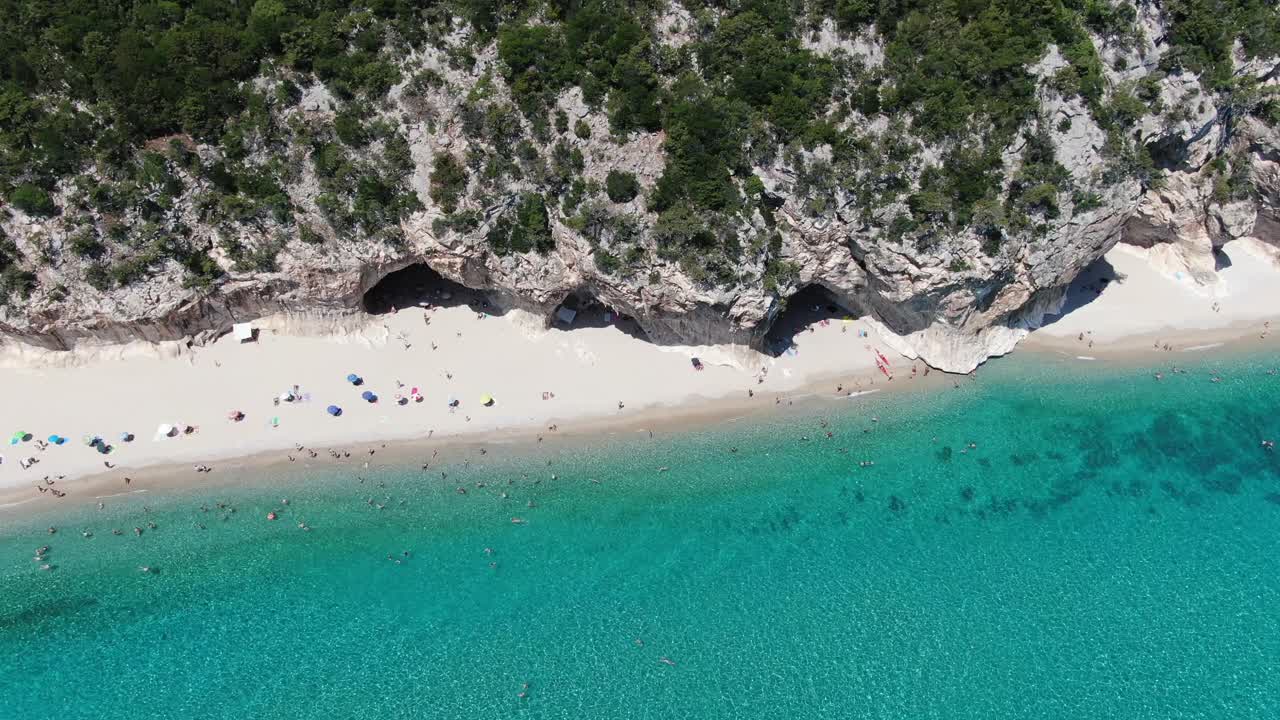 Denne video viser en venstre sideudsigt, der omfatter en krystalklar blå kystlinje med hvide sandstrande og tårnhøje massive klipper. Videoen zoomer tættere ind på grupperne på stranden.