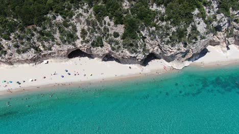 this video follows a left side span view that encompasses a crystal blue coastline with white sandy shores and towering massive cliffs. the video zooms in closer to the groups on the beach