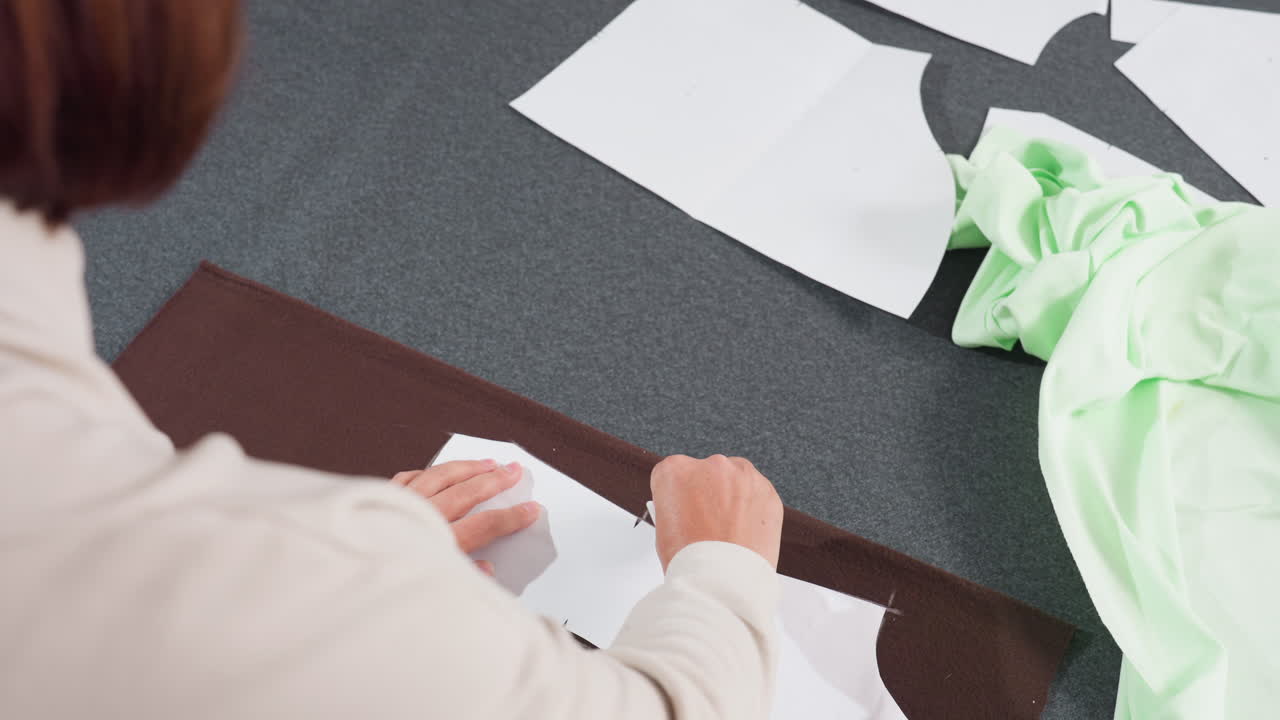 Costumer drawing pattern on white paper sheet placed on dark brown fabric while working on large grey table covered with additional white paper patterns and green fabric inside tailoring workshop