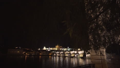 The-Prague-Castle-and-Charles-bridge-over-river-Vltava-in-the-historical-centre-of-Prague,-Czechia,-lit-by-lights-at-night,shot-from-the-other-side-of-the-river,slowly-tilting-through-willow-branches